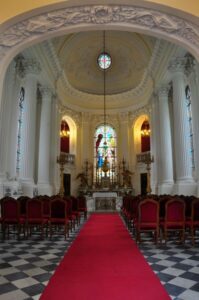 kaplica pałacowa, widok na ołtarz, na środku rozłożony czerwony dywan, po bokach stoją krzesła i widoczne są bogato zdobione kolumny