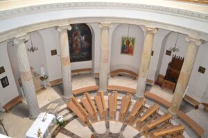 widok okrągłego wnętrza kościoła z poziomu chóru, na ścianach religijne obrazy w centrum widoczne ławki i kolumny.