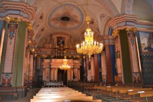 wnętrze kościoła rozświetlone dwoma żyrandolami, bardzo bogato zdobione kolorowymi malowidłami ściany.