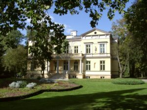 słoneczny dzień, widok na odnowiony pałac w barwach żółto białych otoczony zielonym parkiem z bujną zielenią i kolorowymi roślinami.