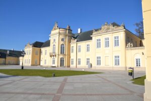 widok na Pałac Potockich z zewnątrz w promieniach wiosennego słońca. Pałac koloru jasno żółtego z białą stolarką, od frontu widoczny dziedziniec z szaro czerwonymi płytami i zielony trawnik